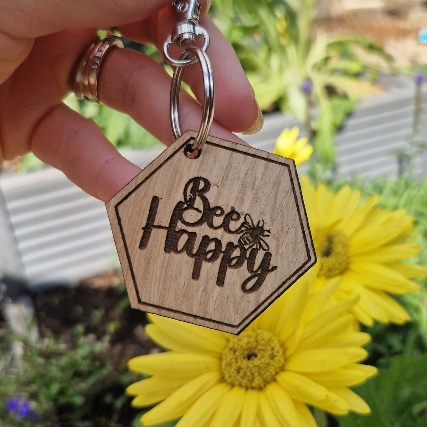 Wooden keychain with 'Bee Happy' text held by a hand with a yellow flower in the background