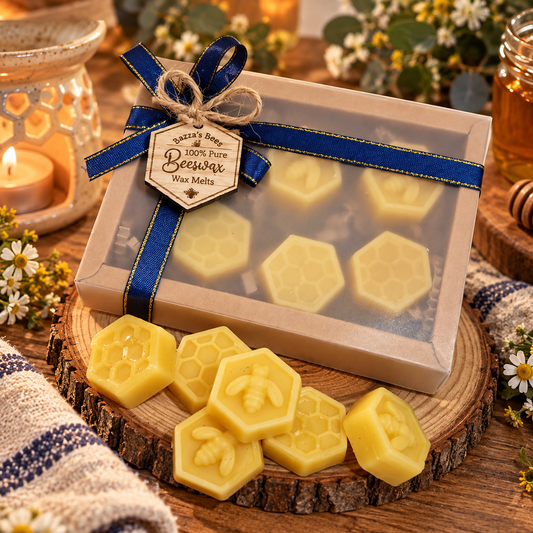 A gift box of handmade beeswax melts and hexagonal beeswax bars, tied with a blue ribbon and hanging tag, displayed on a wooden slice with loose beeswax bars in front. The scene is styled with a lit candle in a glass holder, a jar of honey, and small white flowers on a rustic wooden table.