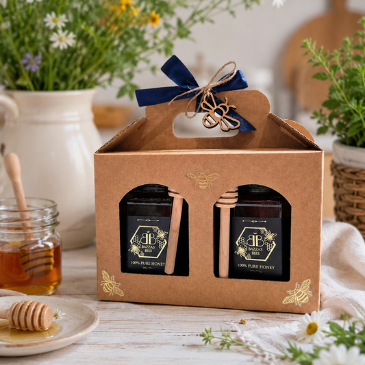 A kraft gift box with a navy ribbon and twine handle, containing two jars of pure honey with wooden spoons, styled on a rustic white table with flowers, a honey dipper, and a jar of honey in the background.